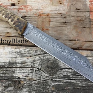 Puede incluir: Un cuchillo grande, hecho a mano, con una hoja de acero estampada y un mango de asta texturizado. La hoja tiene un diseño único y el mango es una mezcla de marrón y beige. El texto "CowboyBlade" es visible.