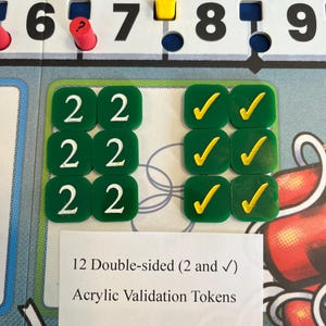 May include: A board game with green acrylic validation tokens. The tokens are double-sided, with the number 2 on one side and a yellow checkmark on the other. The image also shows the numbers 6, 7, 8, and 9.