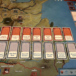 May include: A board game with a map and cards. The cards are arranged in rows, with titles like "Balrog Of Moria" and "A Power Too Great." The game board has a map design with various regions and illustrations. The cards have text and illustrations.