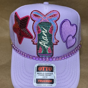 May include: A lavender trucker hat with a mesh back and a curved brim. The front features a star, two hearts, and a watermelon design with the name "Alani". A beaded trim and the "OTTO" logo are also visible.