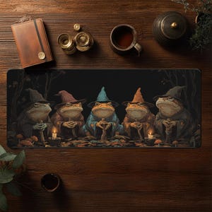 May include: A black desk mat featuring a gathering of five anthropomorphic frogs dressed as wizards. Each frog wears a different colored hat and robe, with candles and mushrooms around them. The mat is rectangular and sits on a wooden surface.