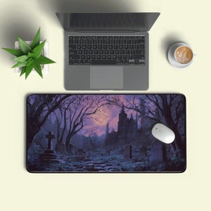 May include: A rectangular mouse pad featuring a gothic graveyard scene with a full moon, bare trees, and a castle in the background. A white mouse and a laptop are on a light beige surface, with a small plant and a cup of coffee.