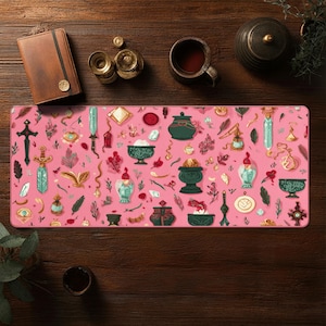 Può includere: Tappetino da scrivania rettangolare rosa con un motivo ripetuto di illustrazioni fantasy. Il design include spade, bottiglie di pozioni, ciotole e altri elementi magici in tonalità di verde, rosso e oro. Un diario in pelle marrone e una tazza di tè sono visibili.