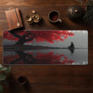 May include: Rectangular desk mat featuring a serene landscape with a red-leafed tree reflected in water. A figure in a boat is visible. The mat is surrounded by a brown leather notebook, brass items, and a cup of coffee.