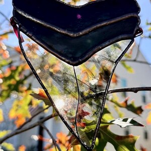 May include: A stained glass shark tooth pendant with a dark blue top section and clear, textured bottom. The edges are outlined in a dark metal, and a pink ribbon is attached for hanging. The background features fall foliage.