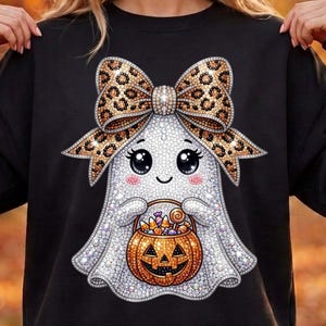 May include: A black long-sleeve sweatshirt featuring a graphic of a white ghost with large black eyes and pink cheeks. The ghost wears a large leopard print bow and holds an orange jack-o'-lantern bucket filled with colorful Halloween candy.