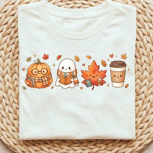 May include: A white t-shirt featuring a graphic print of four cute autumn-themed characters in a row: a pumpkin wearing glasses and a scarf, a ghost reading a book, a maple leaf holding a small cup, and a disposable coffee cup with a face.