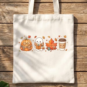 May include: A cream-colored canvas tote bag featuring a central graphic print of a smiling pumpkin wearing glasses and a scarf, a ghost reading a book, an orange maple leaf, and a coffee cup, all surrounded by small autumn leaves and hearts.
