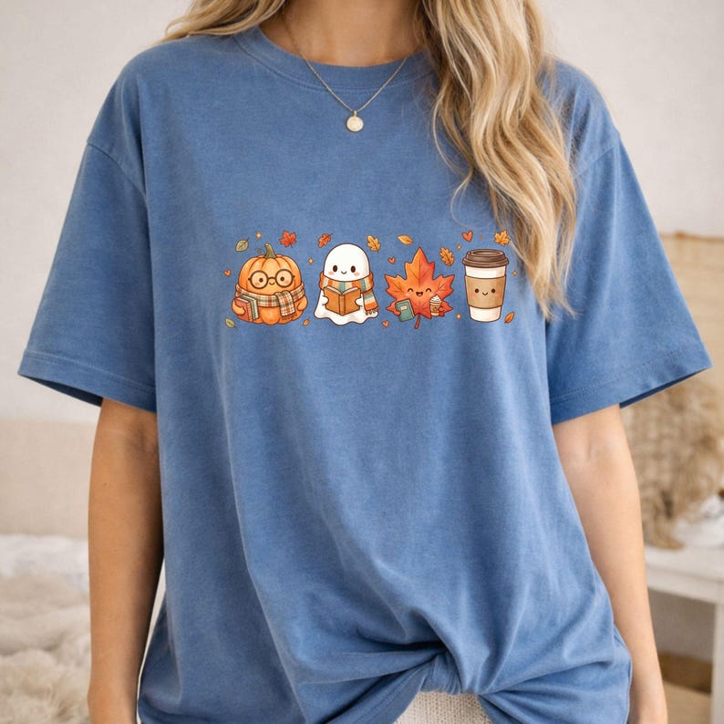 May include: A blue short-sleeve t-shirt featuring a graphic print of a pumpkin wearing glasses and a scarf, a ghost reading a book, an orange maple leaf, and a coffee cup, all surrounded by small scattered autumn leaves.
