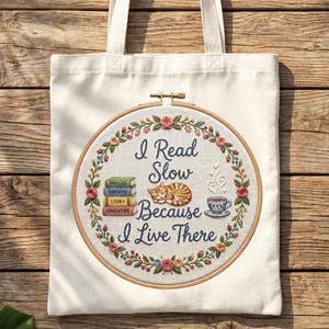 Pode incluir: Saco tote creme com um design bordado circular. O design apresenta o texto "I Read Slow Because I Live There" com uma pilha de livros, um gato dormindo e uma xícara de chá, rodeado por uma coroa de flores.