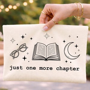 May include: A cream-colored canvas pouch featuring a black line art graphic of an open book, a crescent moon, and eyeglasses, surrounded by small stars. The text "just one more chapter" is printed in a simple black font below the illustrations.