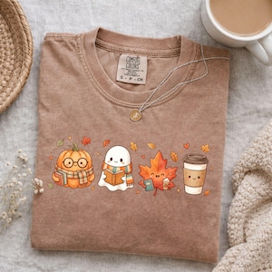 May include: A brown crewneck t-shirt featuring a graphic print of a pumpkin wearing glasses and a scarf, a ghost reading a book, a maple leaf holding a book and coffee, and a coffee cup. The shirt has a Comfort Colors label at the neck.
