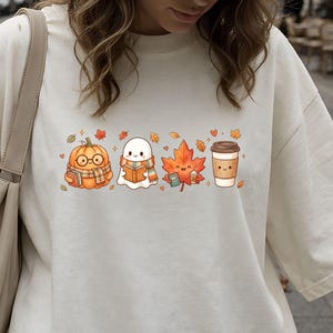 May include: An off-white t-shirt featuring a graphic print of a pumpkin wearing glasses and a scarf, a ghost wearing a scarf, a maple leaf, and a coffee cup, all arranged in a horizontal row with small scattered autumn leaves and hearts.