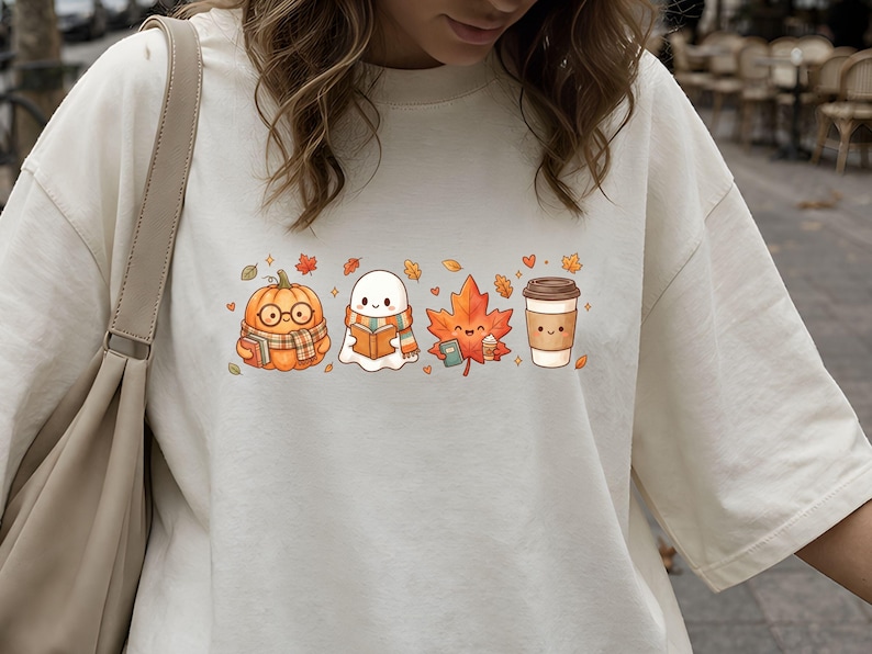 May include: An off-white t-shirt featuring a graphic print of a pumpkin wearing glasses and a scarf, a ghost wearing a scarf, a maple leaf, and a coffee cup, all arranged in a horizontal row with small scattered autumn leaves and hearts.
