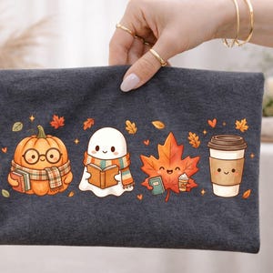 May include: A dark gray fabric pouch featuring a colorful graphic print of a smiling pumpkin wearing glasses and a scarf, a ghost reading a book, a maple leaf holding a small book and coffee cup, and a disposable coffee cup with a smiling face.