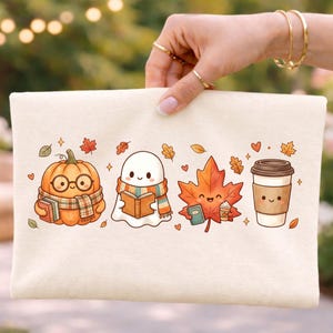 May include: A cream-colored canvas pouch featuring a colorful graphic print of a pumpkin wearing glasses and a scarf, a ghost reading a book, a smiling maple leaf, and a coffee cup, all surrounded by small autumn leaves and hearts.