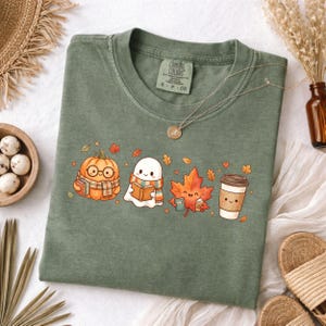 May include: An olive green t-shirt featuring a graphic print of a pumpkin wearing glasses and a scarf, a ghost reading a book, a maple leaf holding a small cup, and a coffee cup. The shirt has a Comfort Colors label at the inner neckline.