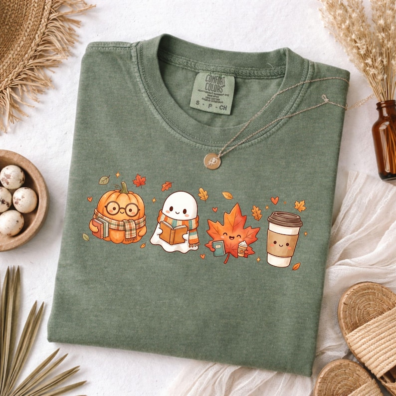 May include: An olive green t-shirt featuring a graphic print of a pumpkin wearing glasses and a scarf, a ghost reading a book, a maple leaf holding a small cup, and a coffee cup. The shirt has a Comfort Colors label at the inner neckline.