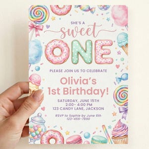 May include: A rectangular birthday invitation card featuring colorful watercolor illustrations of donuts, lollipops, cupcakes, and candy. The text reads: SHE'S A sweet ONE, PLEASE JOIN US TO CELEBRATE, Olivia's 1st Birthday!, SATURDAY, JUNE 15TH, 2:00- 4:00 PM, 123 CANDY LANE, JACKSON, RSVP to Sophie by June 6th, 123-456-7890.