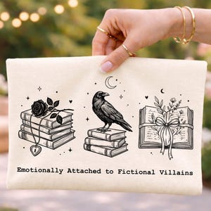 May include: Cream-colored canvas clutch with a black graphic design. The design features stacks of books, a raven, and an open book with floral accents. The text "Emotionally Attached to Fictional Villains" is printed below the illustrations.