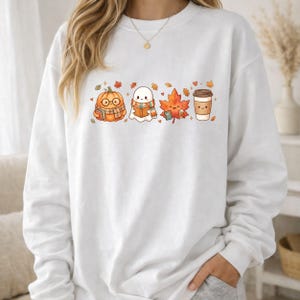 May include: A white crewneck sweatshirt featuring a central graphic print of a cartoon pumpkin wearing glasses and a scarf, a ghost reading a book, a maple leaf, and a coffee cup, all surrounded by small scattered autumn leaves and hearts.