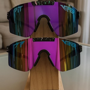 Puede incluir: Dos pares de gafas de sol Viper con lentes espejadas en tonos morados, azules y amarillos. Las gafas tienen monturas negras y la palabra "VIPER" est&aacute; impresa en las lentes. Se exhiben en un soporte de madera.