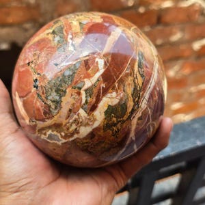 Peut inclure: Une pierre sphérique polie avec un aspect marbré. La pierre présente un mélange de couleurs rouge, marron et crème, créant un motif unique. La sphère est tenue dans une main, mettant en valeur sa taille et sa texture.