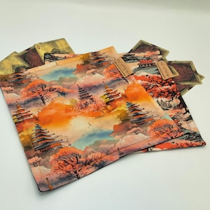 Puede incluir: Varios artículos de tela cuadrados con un diseño de paisaje japonés. El diseño presenta pagodas, árboles y montañas en tonos naranja, rosa y azul. Los artículos de tela están apilados uno encima del otro.