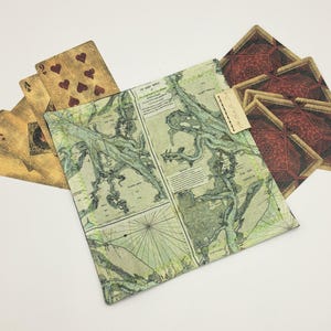 Könnte beinhalten: Ein quadratischer Stoffbeutel mit einem Vintage-Kartenaufdruck in Grün- und Cremetönen, versehen mit einem kleinen rechteckigen Etikett. Der Beutel ist von mehreren Spielkarten mit goldfarbenen Vorderseiten und dunkelroten Rückseiten umgeben.