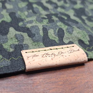 Puede incluir: Primer plano de una tela con estampado de camuflaje en tonos verdes, grises y negros. Una etiqueta rectangular de corcho está adjunta, cosida con hilo verde, y presenta el texto "Ace Hake On Duck".