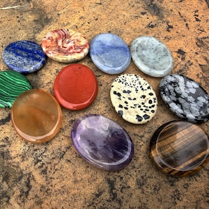 May include: A collection of polished gemstones in various colors and patterns. The stones include lapis lazuli, red jasper, amethyst, and tiger's eye. The stones are oval and round, and are displayed on a brown, textured surface.
