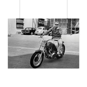 Peut inclure: Photographie en noir et blanc d'un homme sur une moto vintage. La moto a un grand pneu avant et un petit réservoir d'essence. L'homme porte des lunettes de soleil et une chemise boutonnée. Voiture classique en arrière-plan.