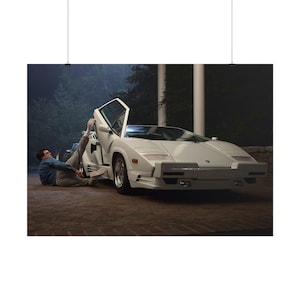 May include: A white Lamborghini Countach, a classic sports car, with its distinctive upward-opening door. The car is parked on a brick surface. A person in a denim jacket and grey trousers is near the car. The scene is at night.