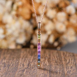 May include: A rose gold pendant necklace featuring a vertical bar design set with a row of multi-colored baguette-cut gemstones. The stones transition in color from light green at the top to pink, purple, blue, and light green at the bottom.