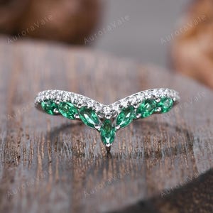May include: A curved, silver-toned ring with a row of small, clear stones along the top and a series of green, marquise-cut stones forming a V-shape. The ring is set against a blurred wooden background.