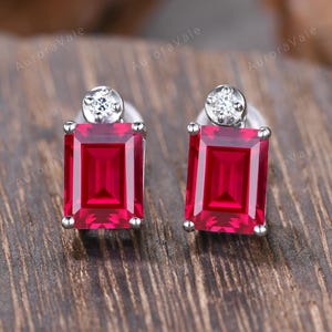 May include: A pair of stud earrings with rectangular, deep red gemstones. Each earring has a small, round, clear stone at the top. The earrings are set in silver metal. The background is a dark wood surface.