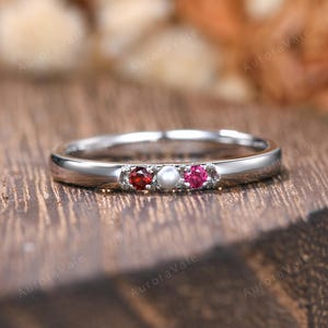 May include: A silver ring with three gemstones: a red ruby, a white pearl, and a pink ruby. The ring is set on a wooden surface, with the words "Aurora Vale" in the background.