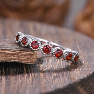 May include: A silver ring with a series of round, vibrant red gemstones set in a scalloped bezel design. The ring is displayed on a textured wooden surface, showcasing the intricate details of the jewelry.