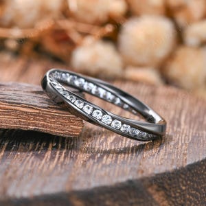 May include: A black metal band ring featuring a channel setting with a continuous row of small, round, clear white stones embedded along the center. The ring has a smooth, polished finish and a slightly twisted or contoured design profile.