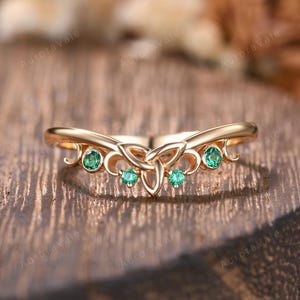 Könnte beinhalten: Ein goldfarbener Ring mit einem keltischen Knotenmuster und fünf runden, grünen Edelsteinen. Der Ring hat eine geschwungene Form und ist vor einem Holzgrund platziert. Das Design beinhaltet dekorative Wirbel.