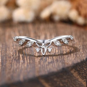 Könnte beinhalten: Ein silberfarbener Ring mit einem Blatt- und Dreifaltigkeitsknoten-Design. Der Ring hat kleine, klare Edelsteine, die in den Blättern und in der Mitte eingesetzt sind. Der Ring ist vor einem verschwommenen Holzgrund gesetzt.