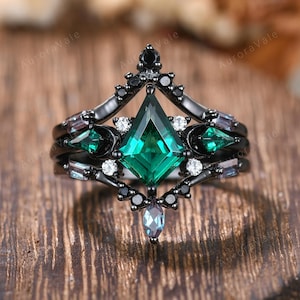 May include: A black ring set with a large, emerald-cut green gemstone at the centre, surrounded by smaller green and white stones. The ring has a unique, layered design with a crown-like top. The background is a wooden surface.