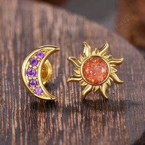 May include: A pair of gold-colored stud earrings. One earring is a crescent moon shape with purple gemstones. The other earring is a sunburst design with an orange center. The earrings are displayed on a wooden surface.