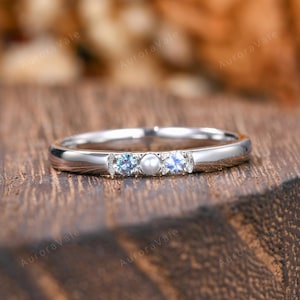 May include: A silver-toned band ring featuring a central white pearl flanked by two small, round, iridescent blue-toned gemstones. The metal band has a smooth, polished finish and a slim profile, showcasing a delicate and symmetrical arrangement.