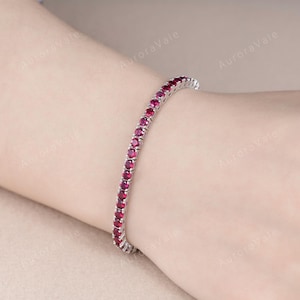 May include: A silver bracelet with a row of round, vibrant red gemstones. The gemstones are closely set together, creating a continuous line of color. The bracelet is shown on a person's wrist.