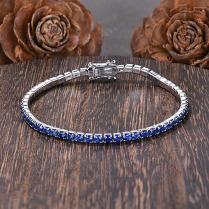 May include: A silver bracelet with a row of small, vibrant blue gemstones. The bracelet is set against a dark wooden surface, with the clasp visible. The gemstones are a deep, saturated blue color.