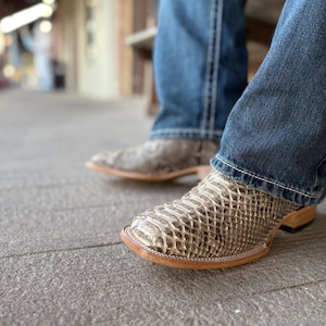 Könnte beinhalten: Nahaufnahme von Cowboy-Stiefeln mit Schlangenmuster. Die Stiefel sind hellbraun und beige mit quadratischer Spitze und Ledersohle. Die Person trägt eine blaue Jeans.