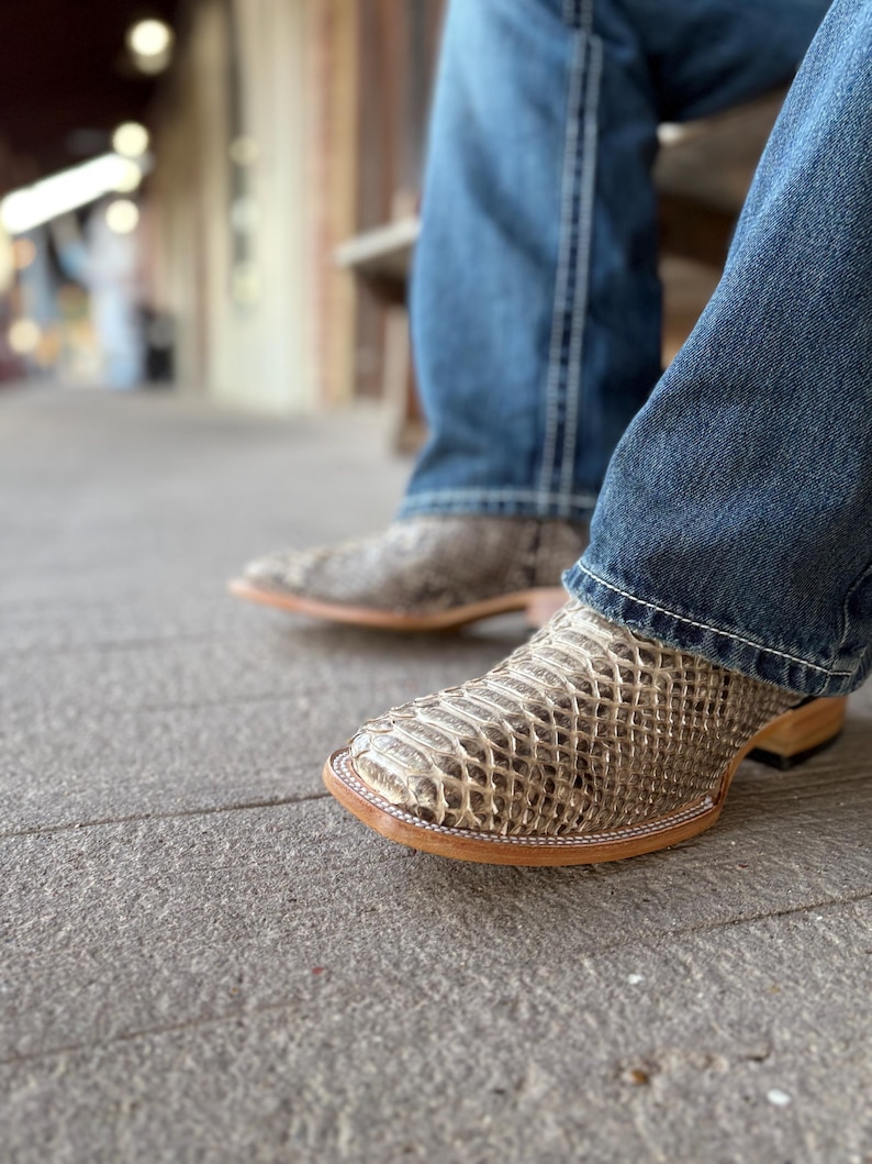 Peut inclure: Gros plan sur une paire de bottes de cowboy &agrave; motif peau de serpent. Les bottes sont marron clair et beige avec un bout carr&eacute; et une semelle en cuir. La personne porte un jean bleu.