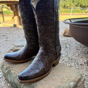 Puede incluir: Un par de botas de vaquero de cuero marrón con intrincados diseños florales. Las botas tienen punta cuadrada, tacón apilado y caña alta. Se exhiben sobre una piedra, mostrando su detallada artesanía.