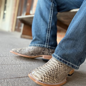 Peut inclure: Gros plan sur une paire de bottes de cowboy &agrave; motif peau de serpent. Les bottes ont un bout carr&eacute; et une semelle marron clair. La personne porte un jean bleu.